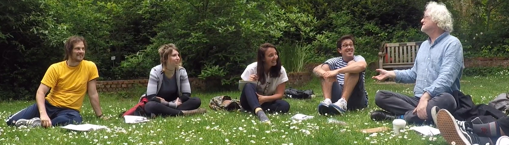 Group of students sit outside in a circle looking at professor, who is speaking and gesturing with his hand.