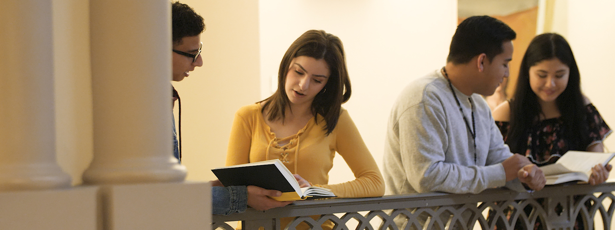 Group of four people discuss open books on balcony railing near marble columns.