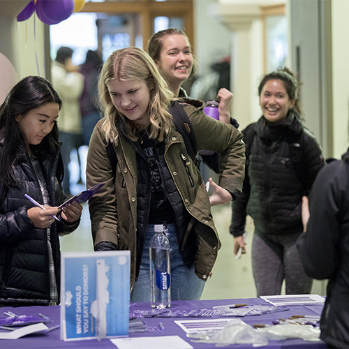 Smiling people approach a purple table with papers on it.