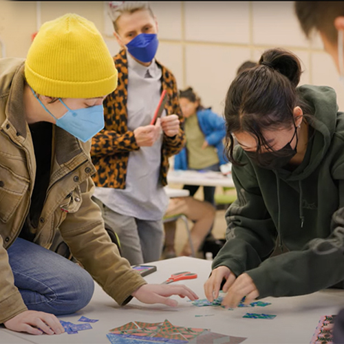 Students around a table with colorful shapes, collaborating on an art project