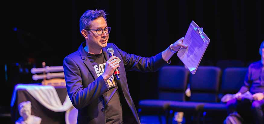 Alex Minami speaking to an audience, with a microphone in his hand. 