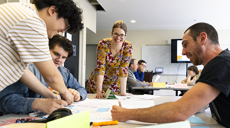 Three students drawing on a large sheet of paper at a table as their professor looks on.