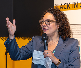 Ralina Joseph holding a microphone and speaking, with an Interrupting Privilege display panel behind her, at the Northwest African American Museum.