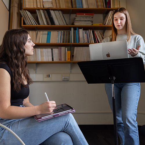 Student standing and speaking while another student sits in front of her, taking notes.