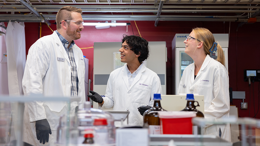 Nick Riley talking with two students in his lab, all of them in white lab coats.